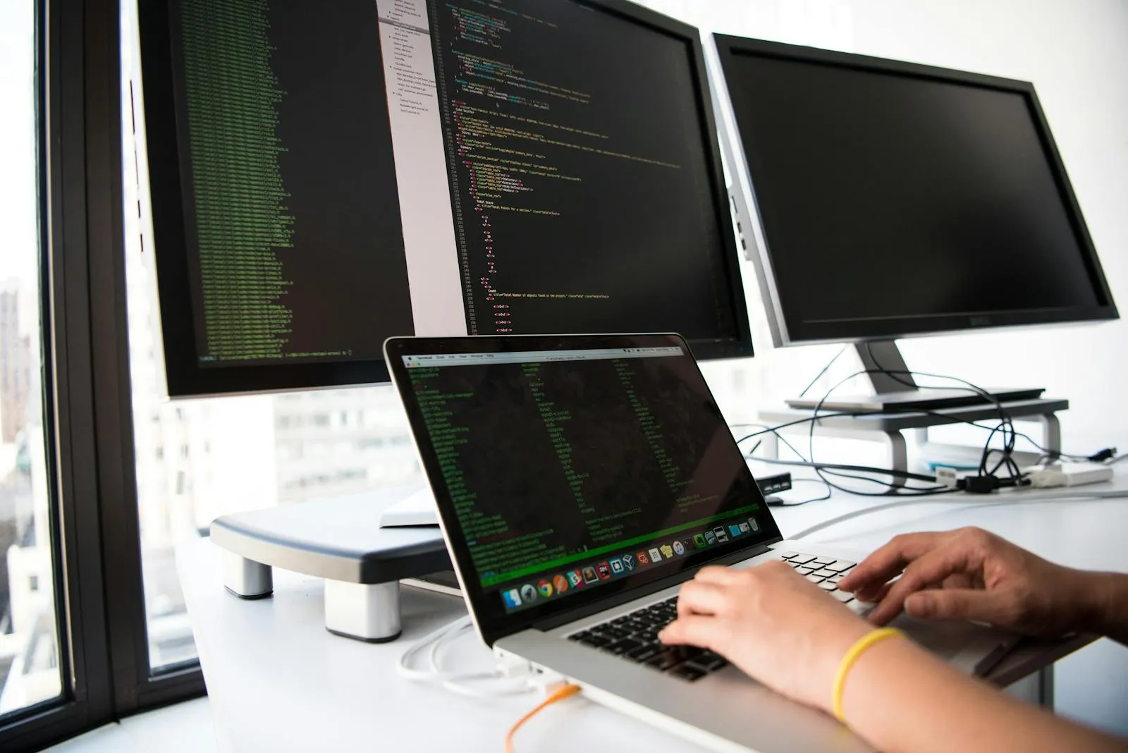 Entwicklungsarbeitsplatz mit Laptop, mehreren Monitoren und aktivem Coding-Kontext.